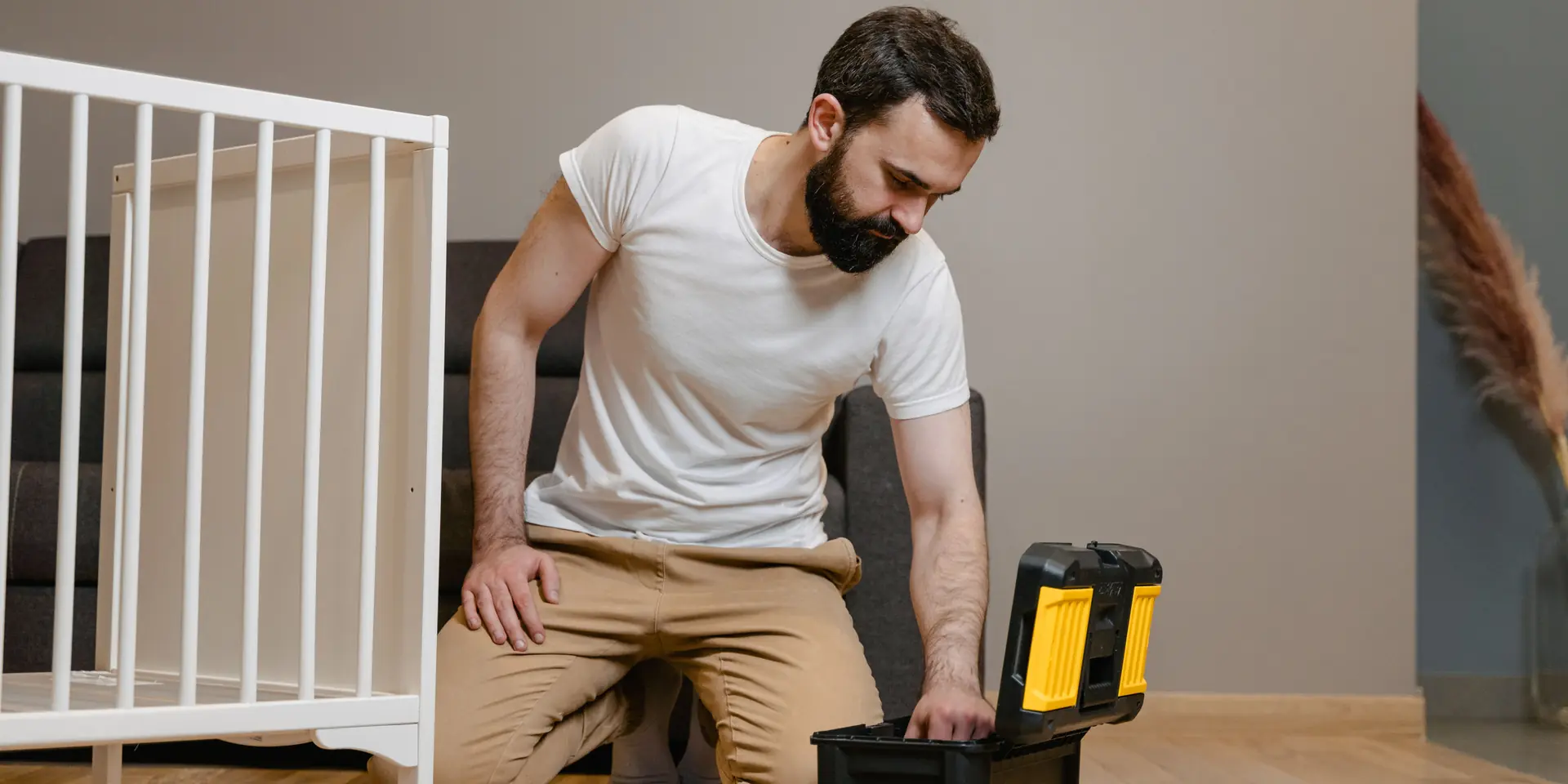 A handyman kneeling by an open toolbox in a living room, beside a cot he is putting together.
