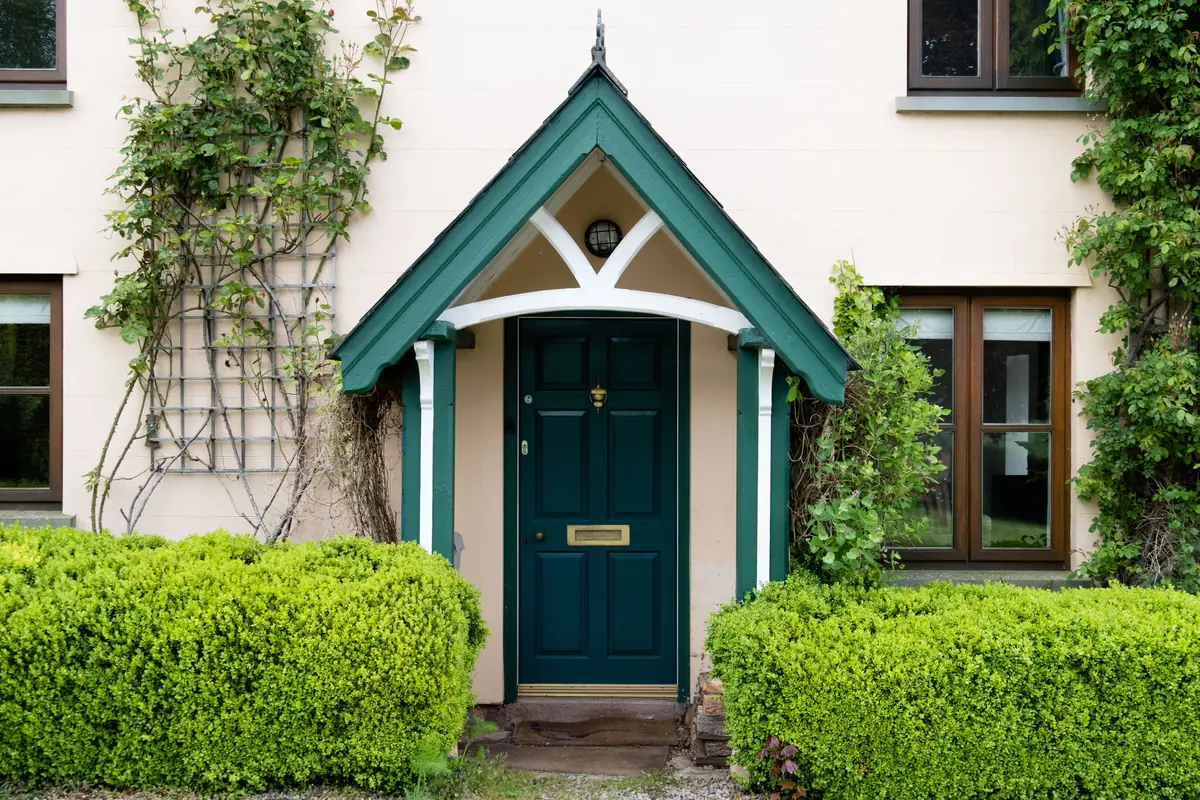 A green front door with a brass letterbox under a gabled porch, between two trimmed hedges.